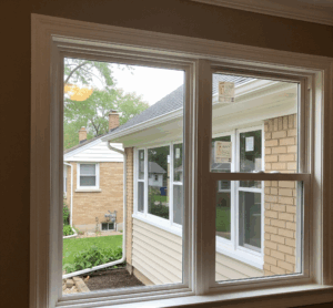 A new, white-framed window is installed in a house, looking out onto a neighboring brick home and a lush green yard by Midwest Windows, Siding & Doors