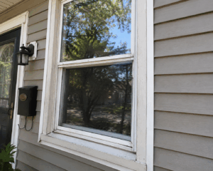 Close-up of an older drafty window with white trim, beige siding, and a black front door on a Chicago residential home.